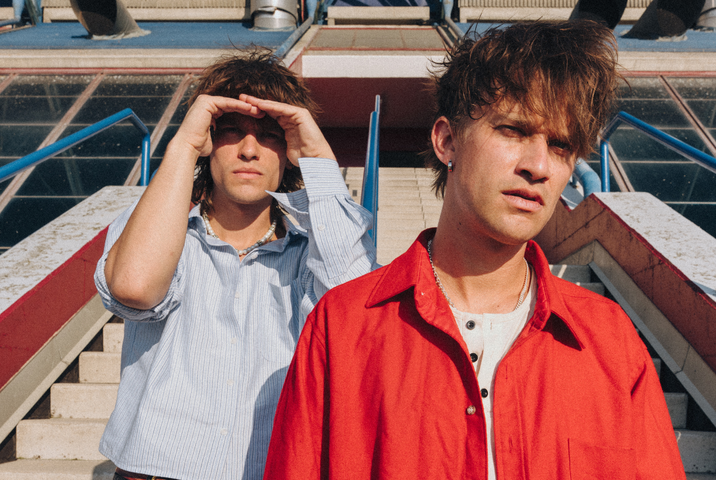Two people one with a button down shirt pn and another with a red back standing on steps.